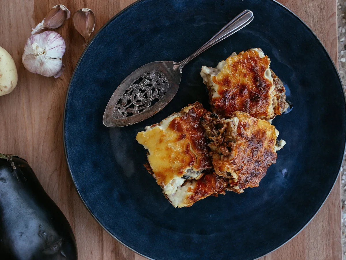 En gyldenbagt aubergine-ret med en gyldenbrun overflade på en mørkeblå tallerken, med en aubergine og hvidløg ved siden af