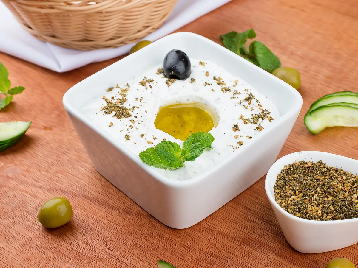 En firkantet hvid skål med cremet yoghurtdyppelse med olivenolie, mynte og krydderurter, ved siden af oliven og agurkeskiver