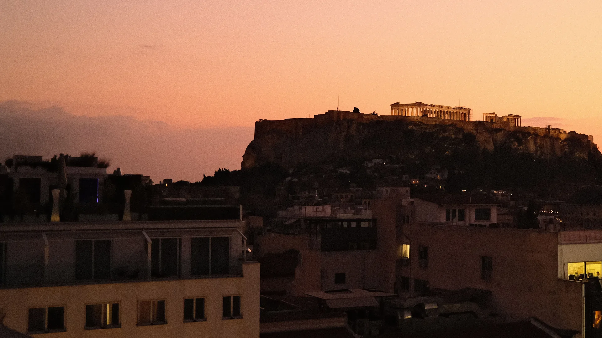 Skumringen over Athen set fra en tagterrasse: Parthenon oplyst på Akropolis-klippen over byens flade tage under en orange himmel
