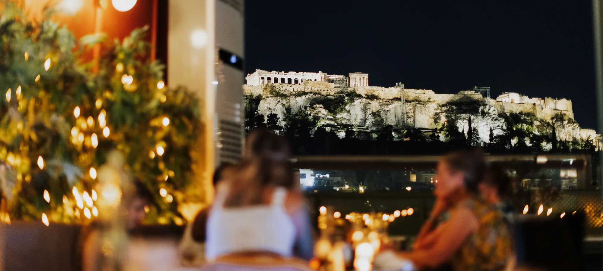 Spisende gæster ved et tagbord oplyst af lyskæder, med den oplyste Akropolis og Parthenon på bakken bag den mørke himmel